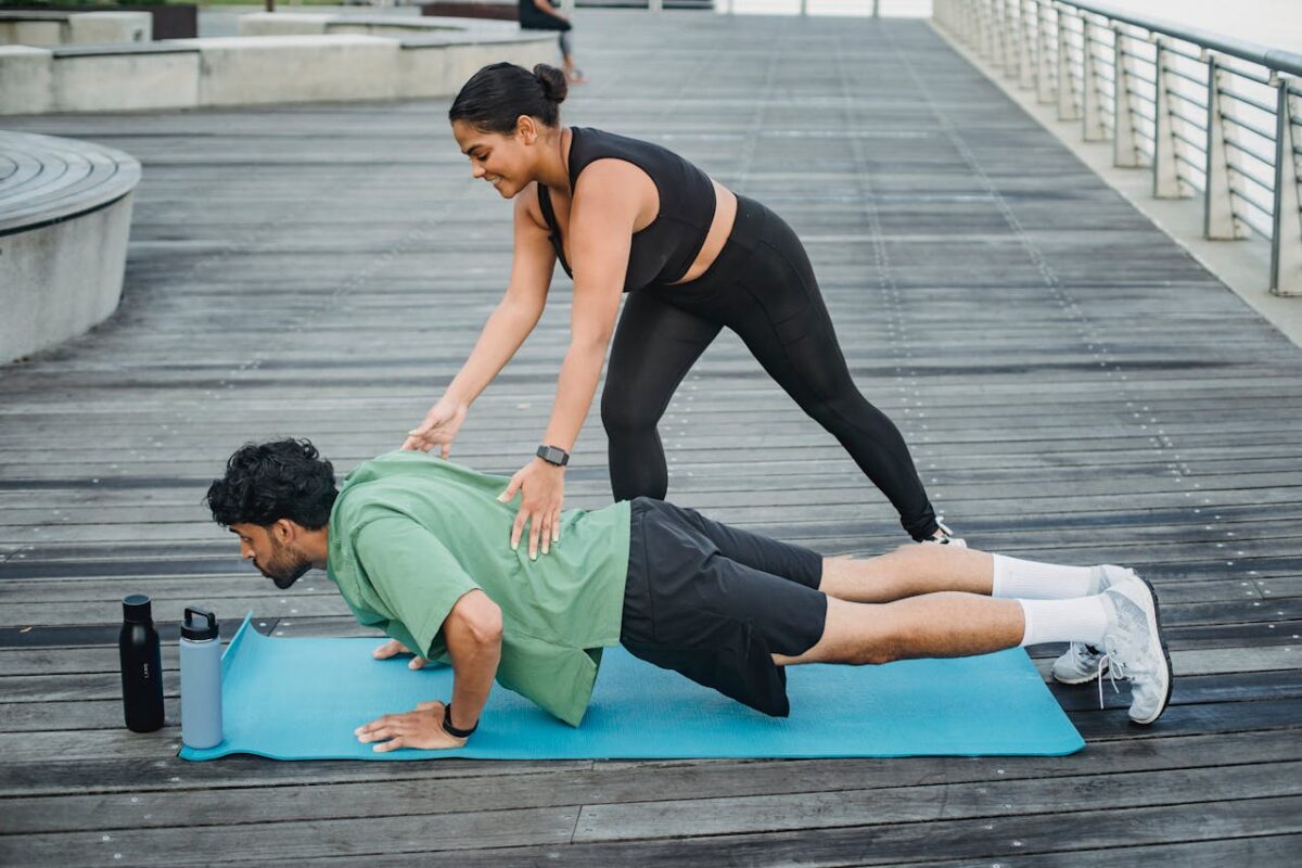 Man doing push-ups with assistance from a trainer on a boardwalk during summer.