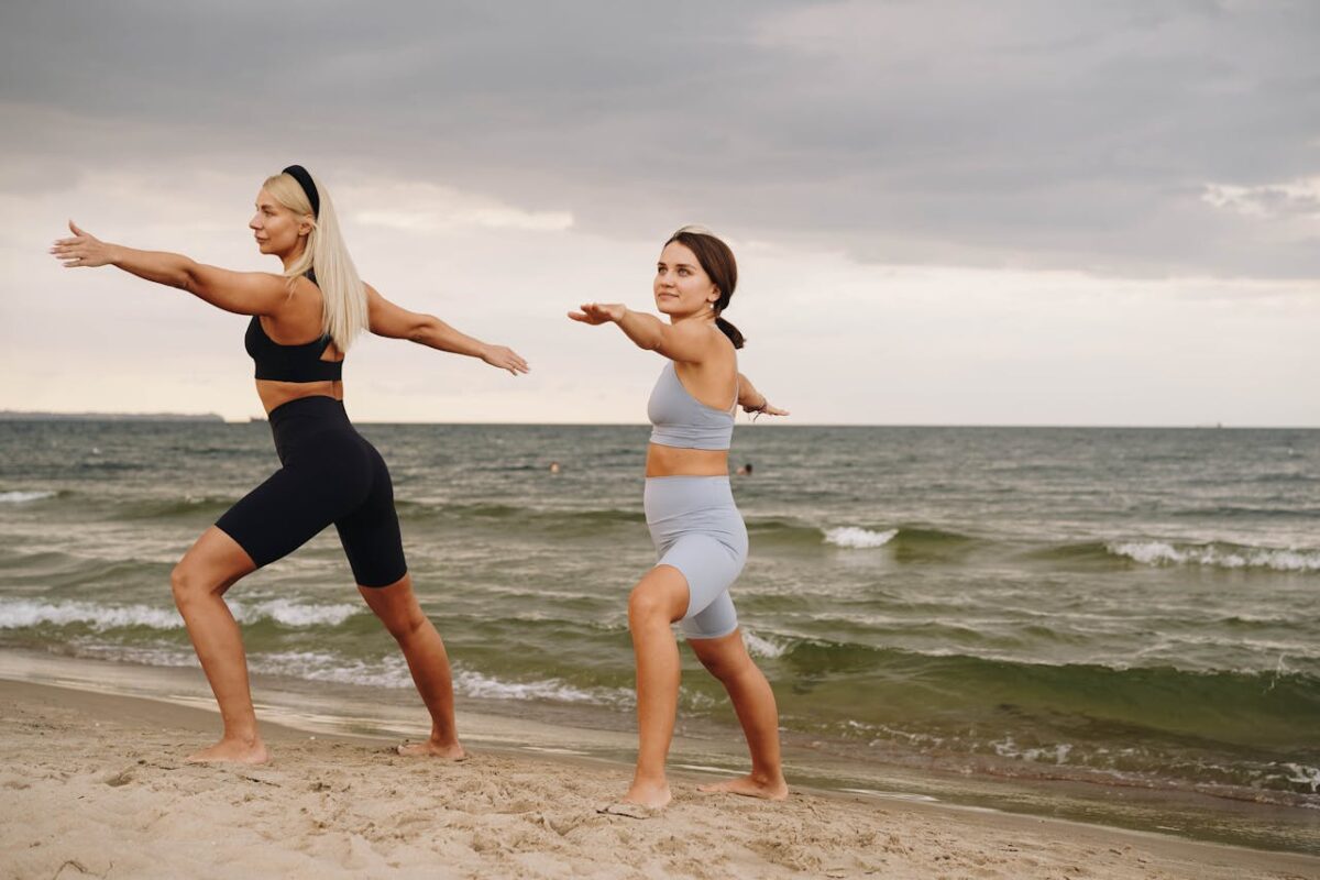 Two women practicing yoga on a sandy beach by the sea, promoting fitness and mindfulness.